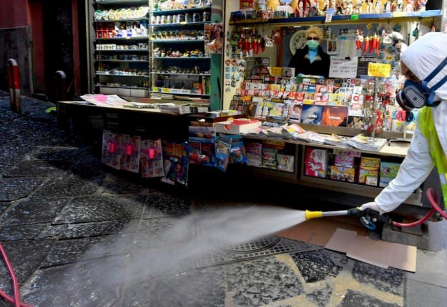 Una persona con tuta protettiva spruzza acqua sul marciapiede fuori da un negozio, presumibilmente per igienizzare.