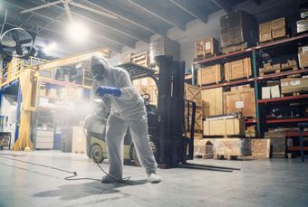 Persona in tuta protettiva che spruzza disinfettante in un magazzino con scatole impilate e un carrello elevatore.