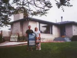 Two people stand in front of a yellow house with a real estate sign.