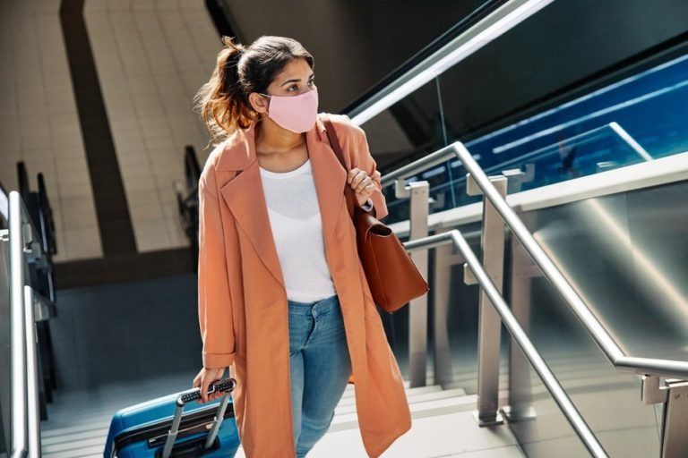 Uma mulher com máscara e casaco rosa, carregando bagagem, sobe uma escada rolante em um prédio