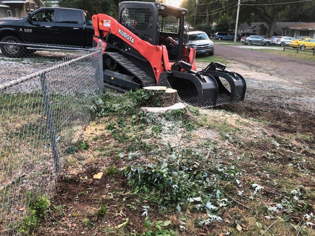 Skid steer with bucket removing a tree stump next to a chain-link fence.