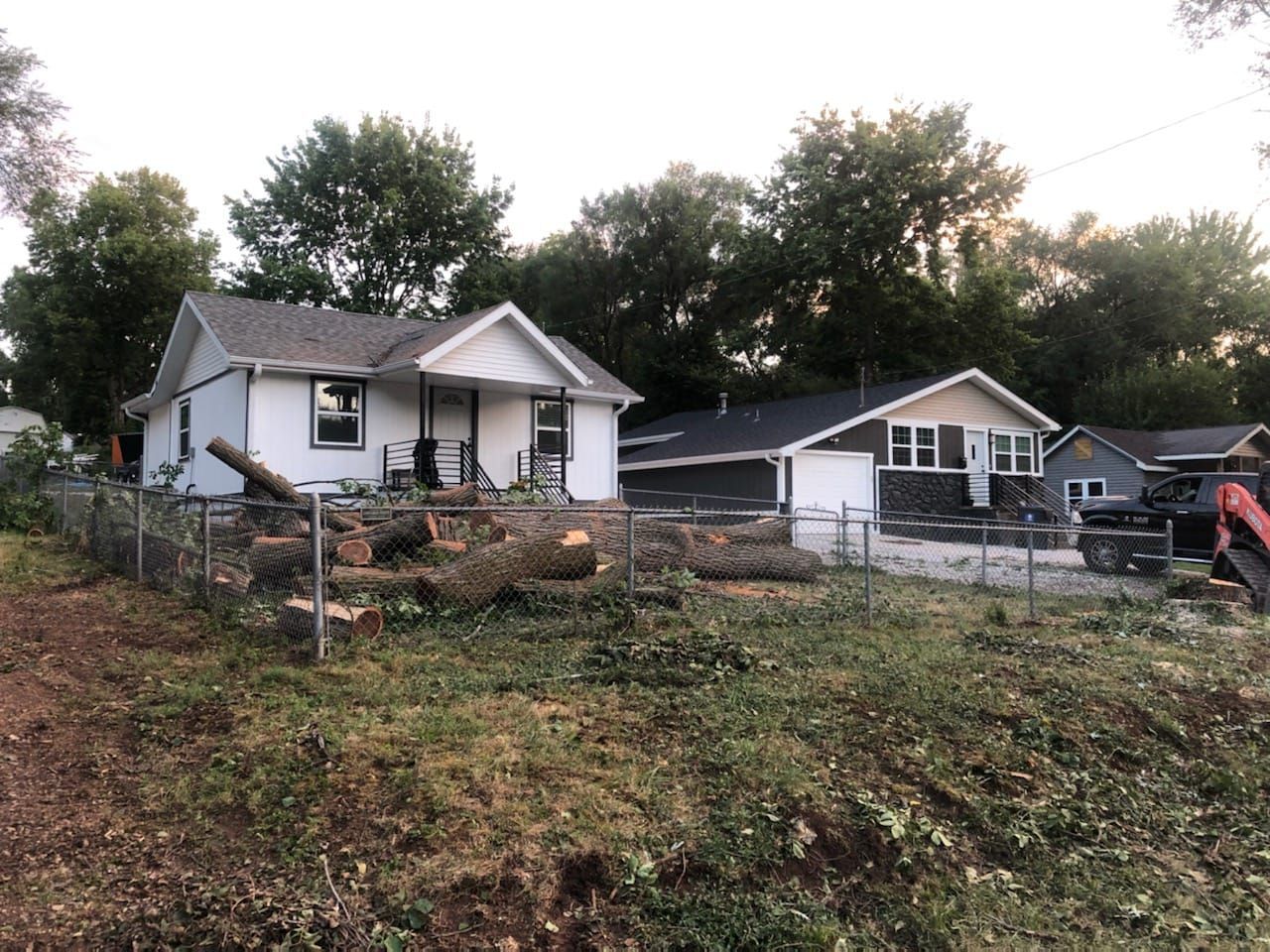 Homes after tree removal; logs in the yard, fence, and a small excavator.