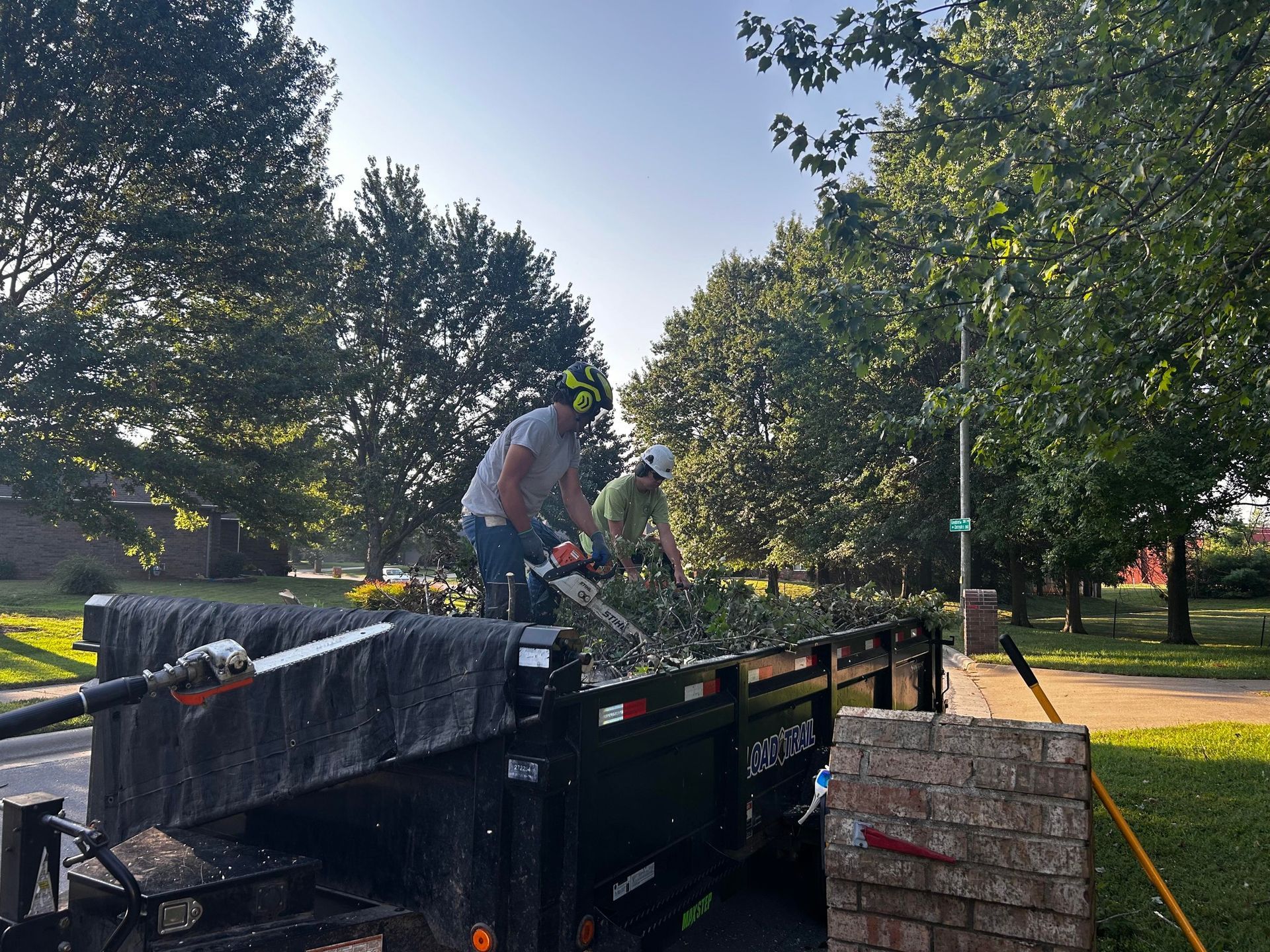 Two people loading tree branches into a dark trailer. They are in a sunny, residential area.