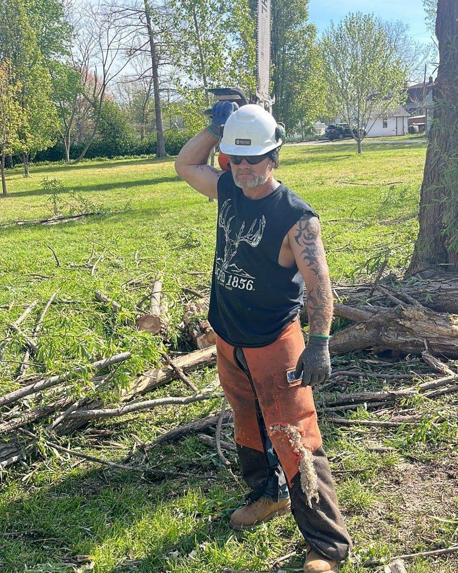 Man wearing safety gear holding a chainsaw outdoors, near cut tree branches and grass.