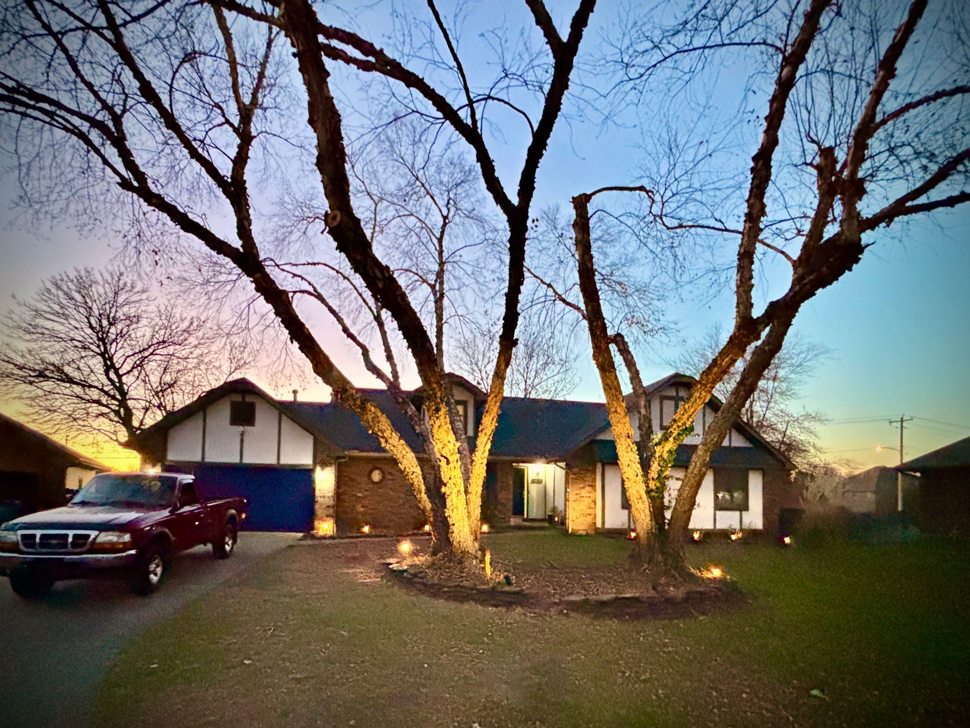 House at dusk, lit trees frame entrance. Red truck in driveway. Blue garage door.