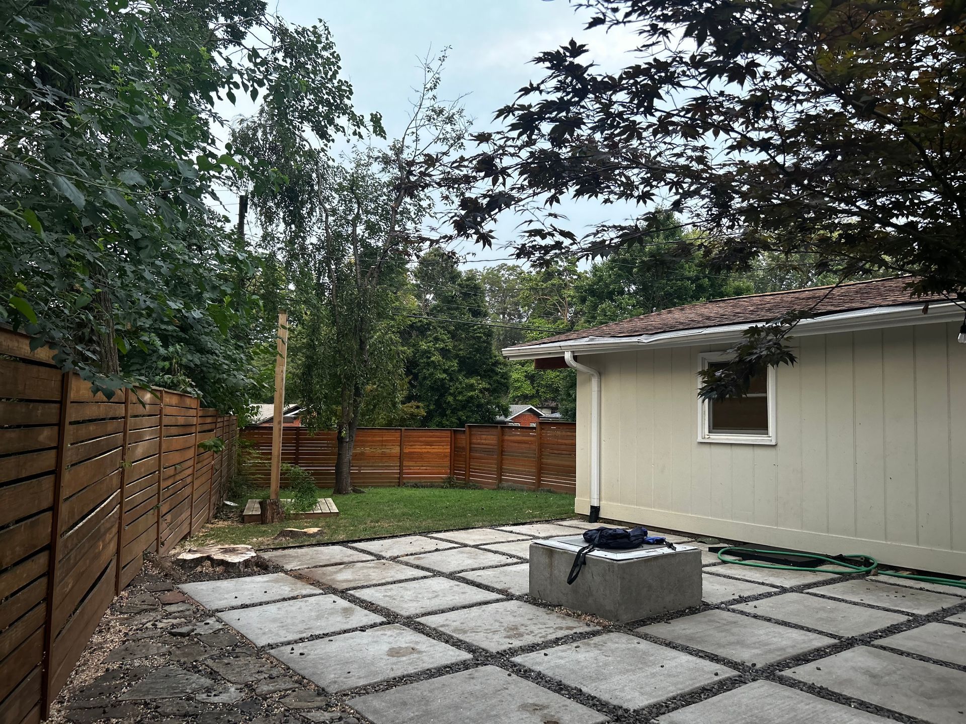 Backyard with stone patio, fire pit, wooden fence, and light-colored house. Trees in the background.
