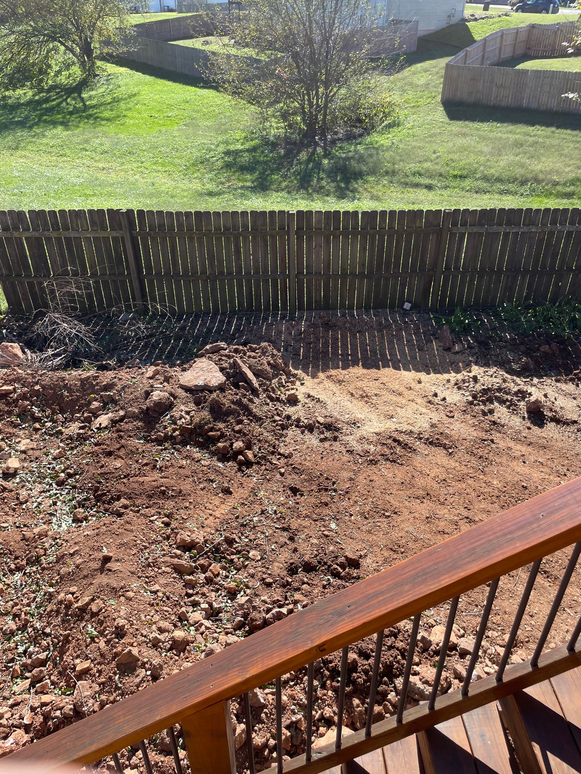 Backyard view from a deck. Brown dirt pile in the foreground, wooden fence, and green grass on a slight incline.