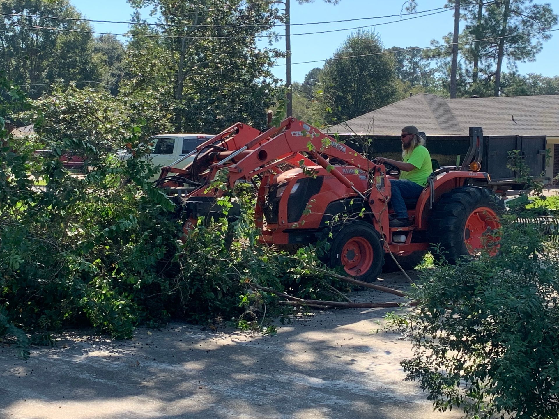Man operating an orange tractor removing brush; parked car and building in background on a sunny day.