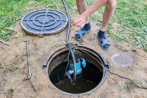A person in sandals lowers a submersible blue pump into an open underground drainage pit in a grassy yard.