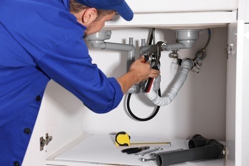 A plumber in a blue uniform uses pliers to repair the pipes underneath a kitchen sink.