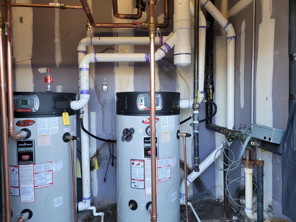Two large grey water heaters side-by-side in a utility room, connected by white PVC venting and copper piping.