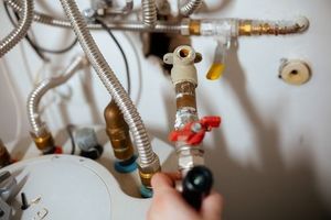 A hand holds a brass pipe fitting with a red lever valve connected to plumbing lines above a water heater tank.
