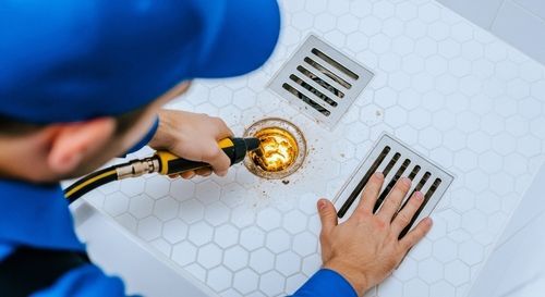 A worker in a blue uniform uses a cleaning tool on a shower drain in a white, hexagonal-tiled floor.
