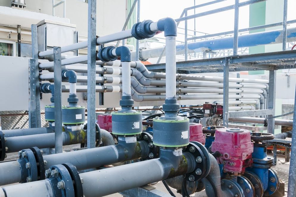Industrial piping system with multiple grey pipes, valves, and flow meters at an outdoor plant facility.