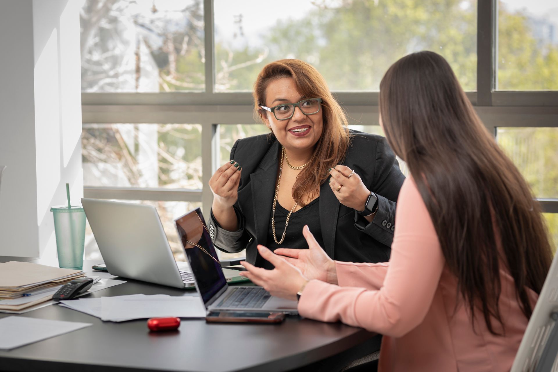 Two women are sitting at a table with laptops and talking to each other.