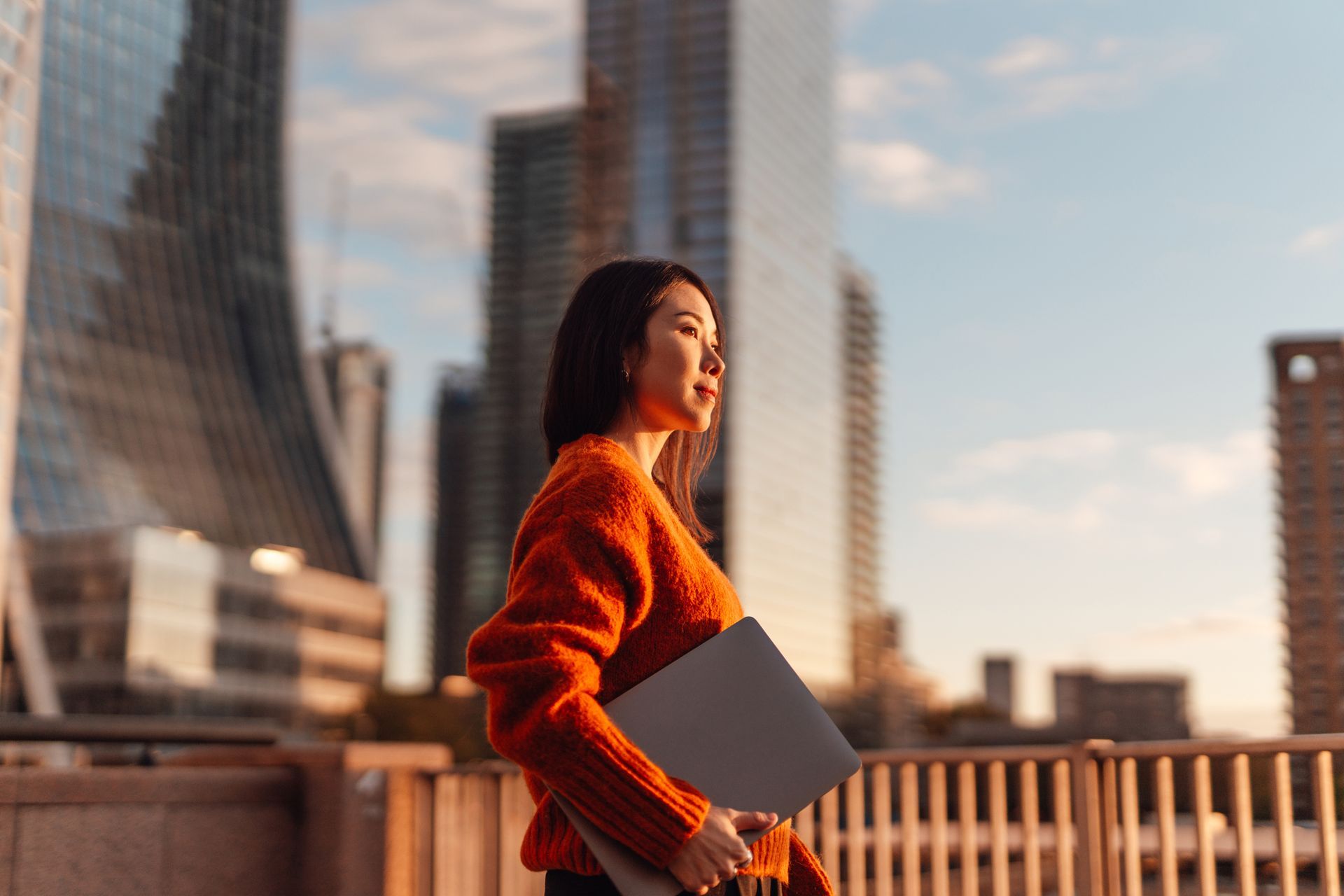A woman in an orange sweater is holding a laptop in front of a city skyline.
