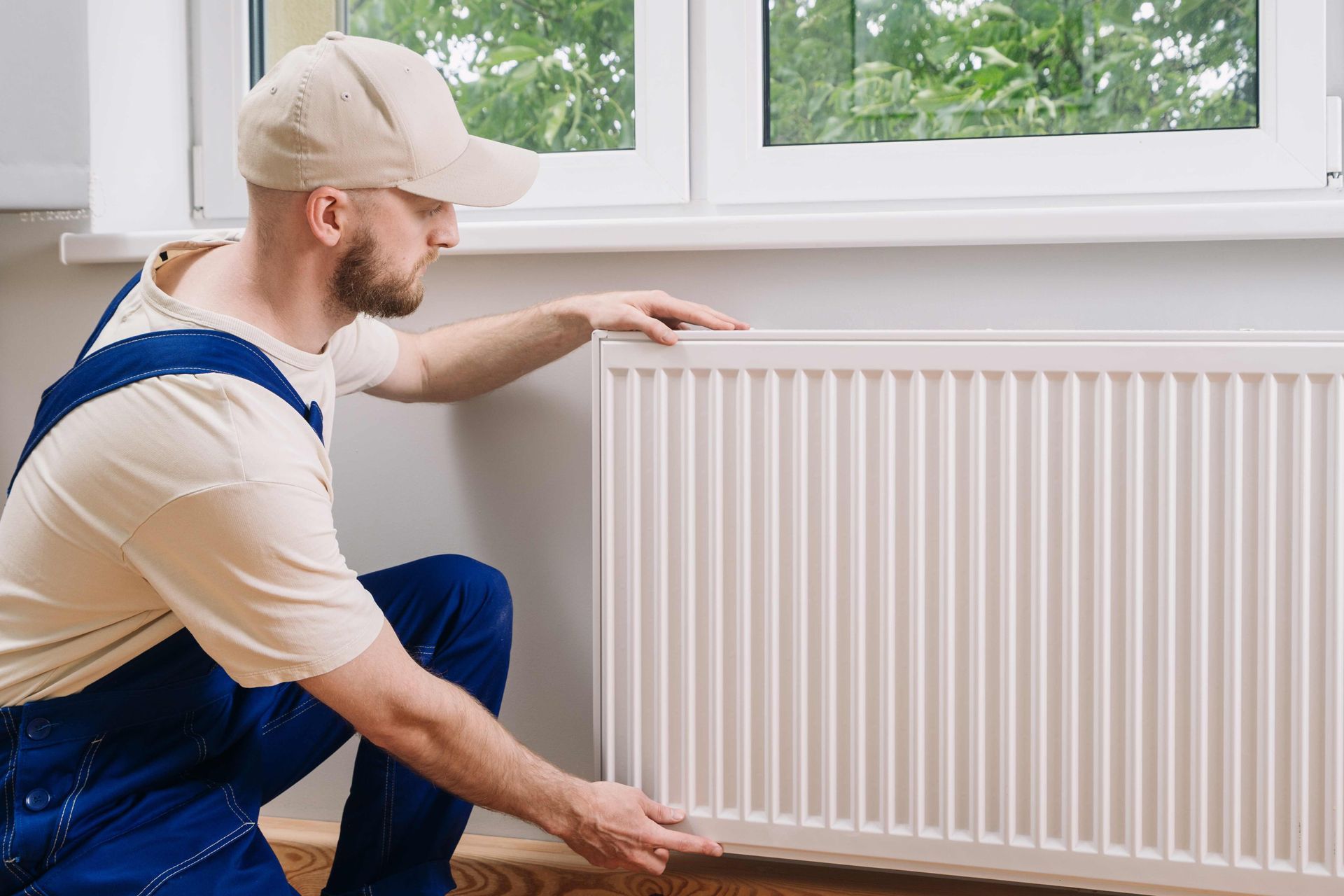 A technician is adjusting a white wall radiator beneath a window during home heating service