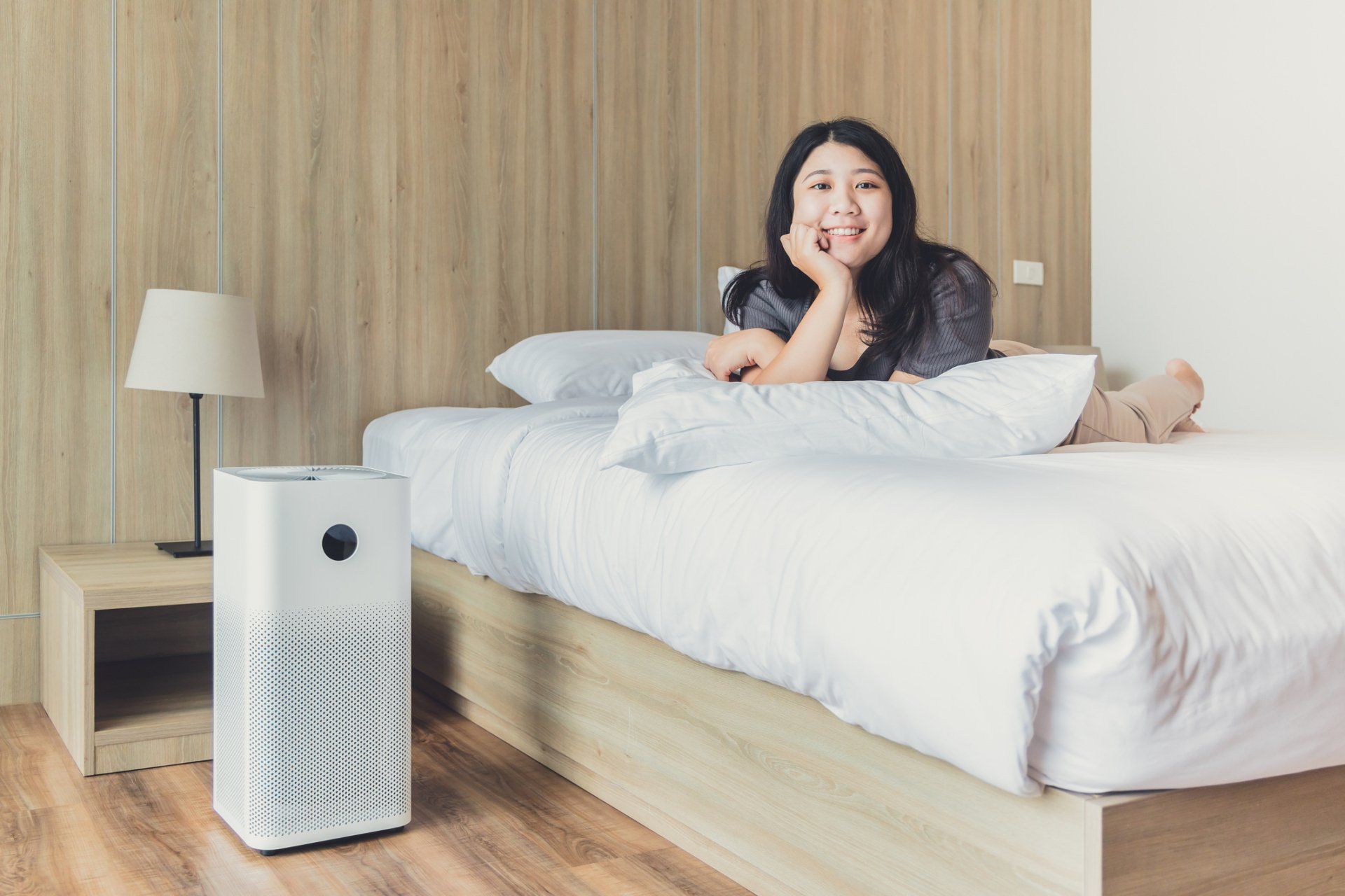 Woman lying on a bed with an air purifier, smiling, light-colored bedroom.