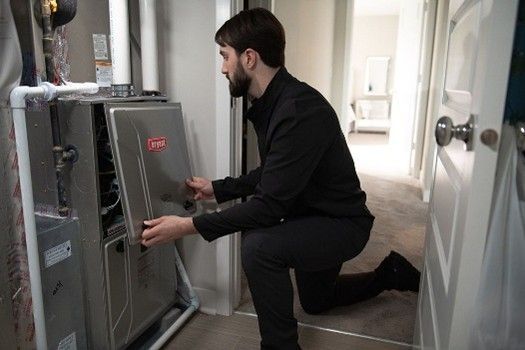 A person kneels in a utility closet, opening the metal service panel of a Bryant HVAC furnace unit.