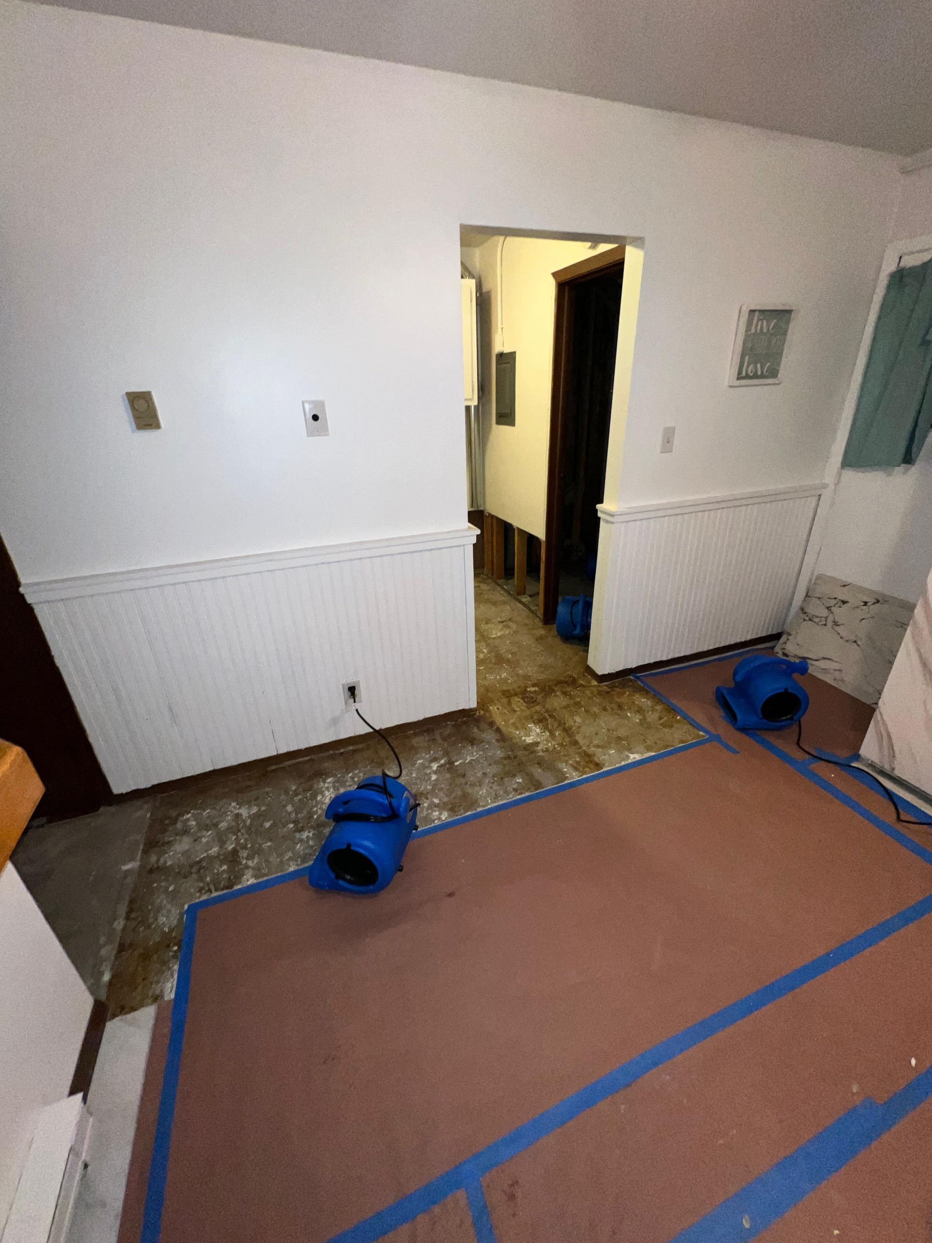 Room under renovation with cardboard-covered floor, blue tape, and doorway in a white interior.