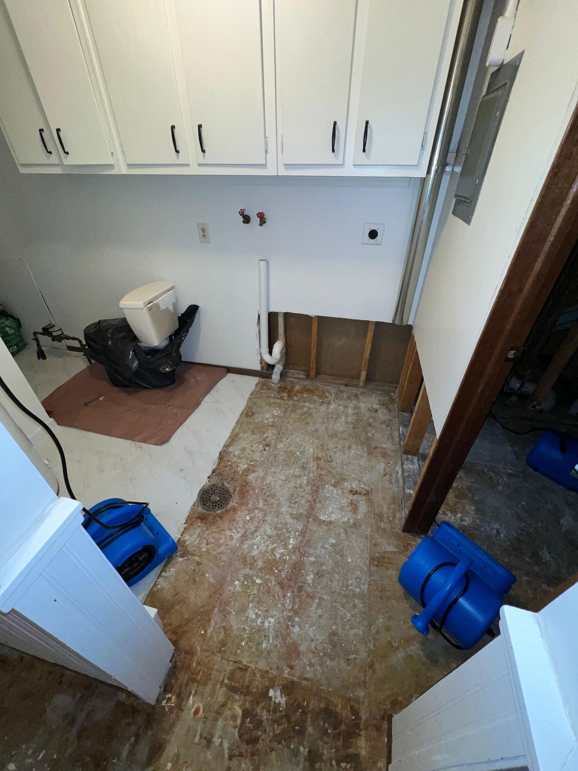 Partially gutted laundry room with torn-up floor, cabinets, and cleaning tools on the left.