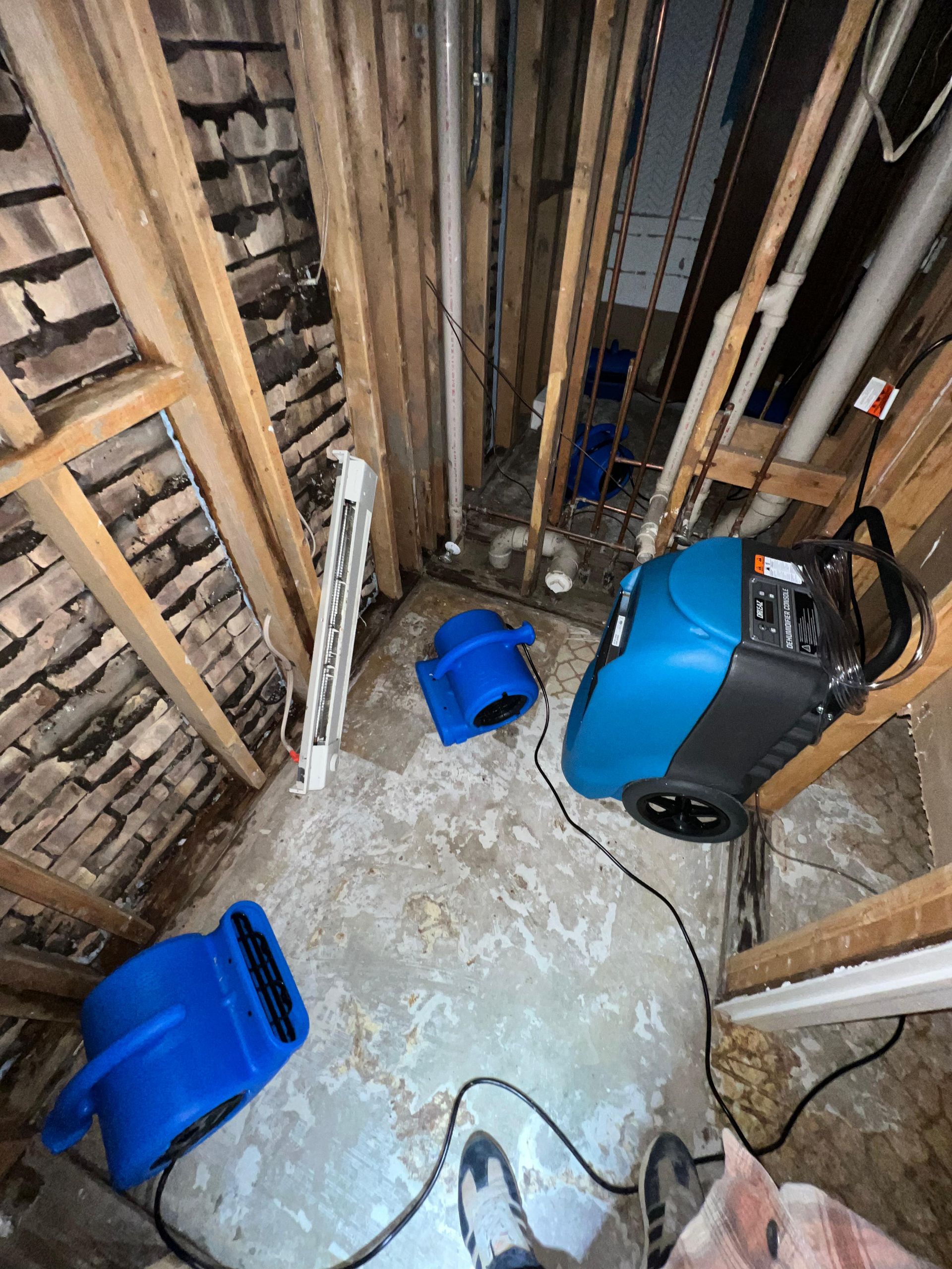 Flood-damaged crawl space with wet floor, exposed studs, blue buckets, and a large fan/dryer.