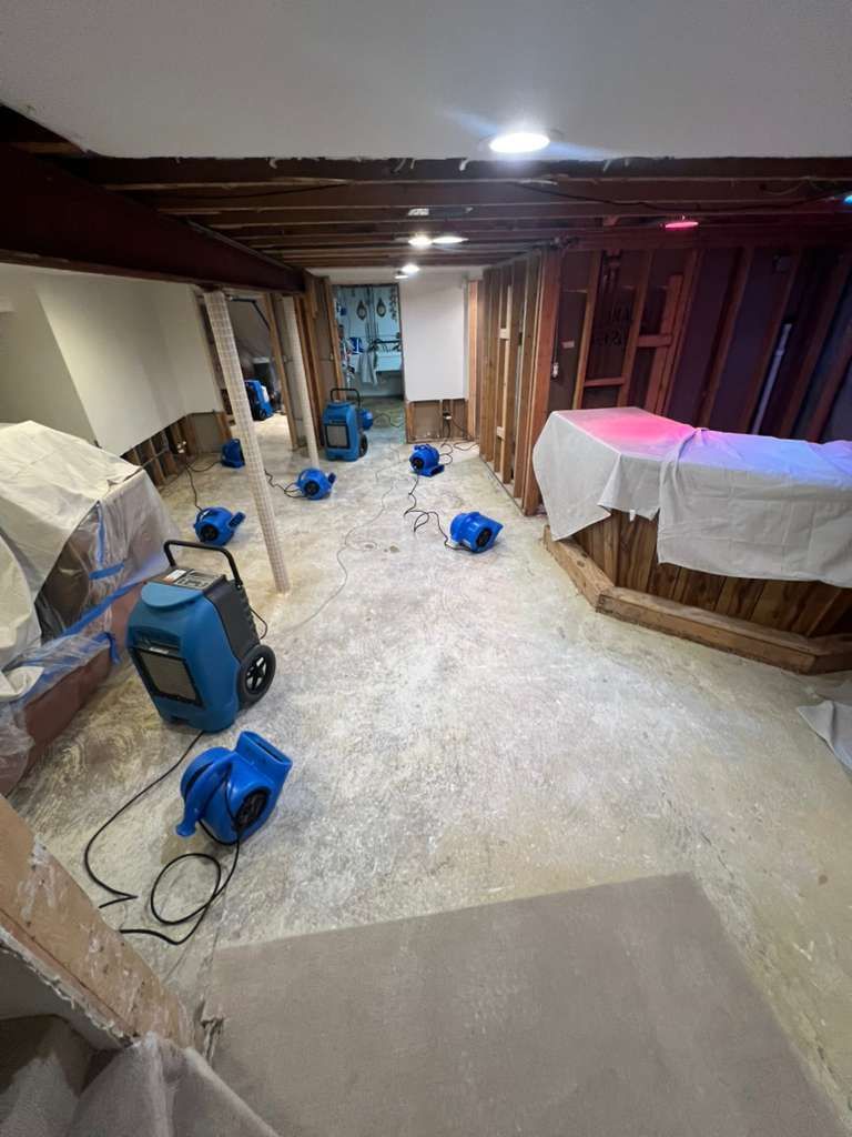 Unfinished basement with exposed beams, tools, blue fans, and covered furniture amid renovation debris