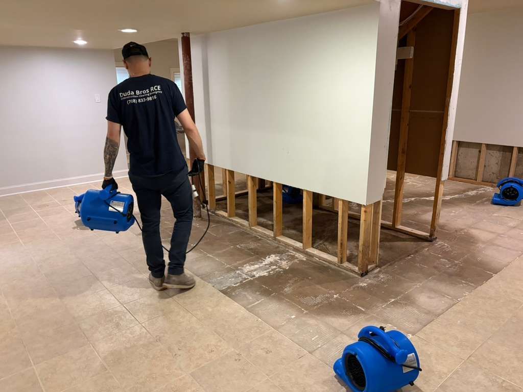 Worker carrying a blue fan in a room under renovation with exposed wall studs and drop cloths.