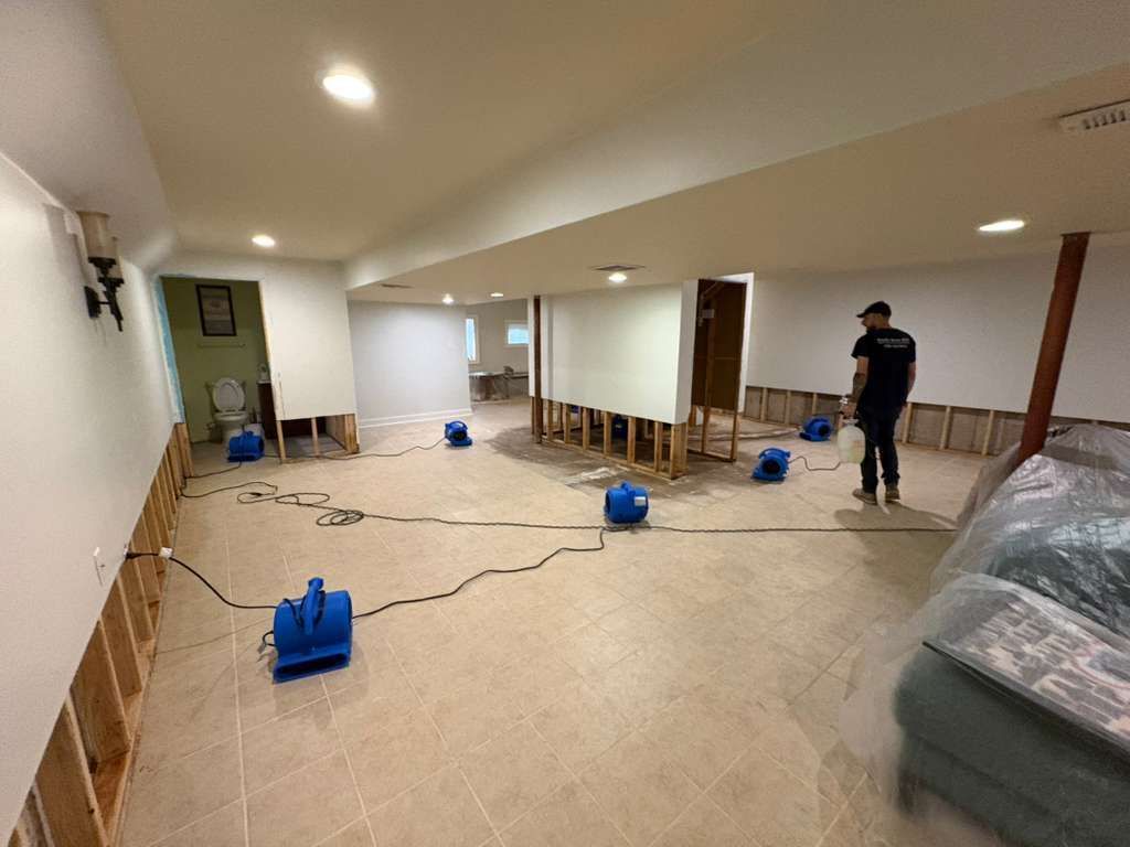 Basement under renovation with a worker, blue fans, plastic-covered furniture, and cords across the floor.