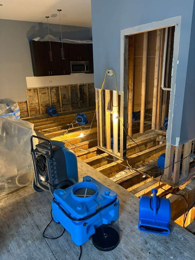 Flood-damaged room with exposed studs, wet flooring, blue cleaning bins, and restoration equipment.