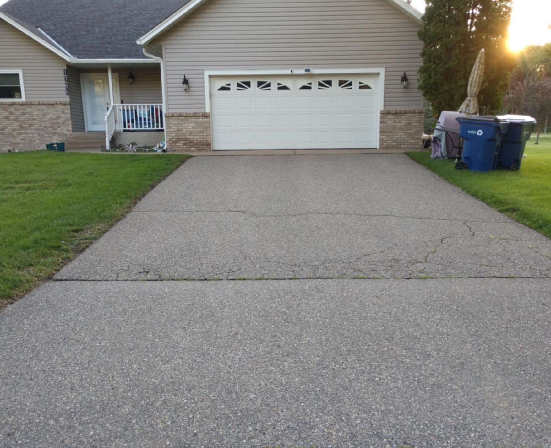 Driveway leading to a house with a white garage door. The driveway is gray and cracked, and the house is tan with a green lawn.