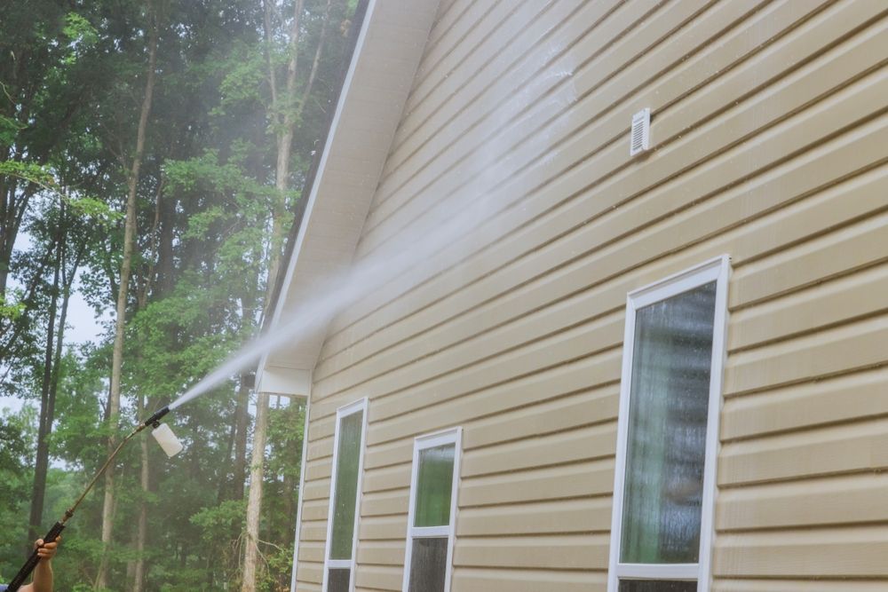 A person power washing the beige siding of a house, creating a stream of water in a wooded setting.