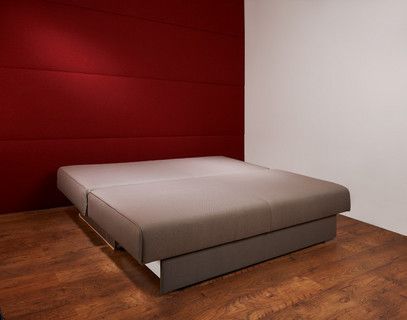 In einem Raum mit roter Wand steht ein ausgezogenes Schlafsofa auf einem Holzboden.
