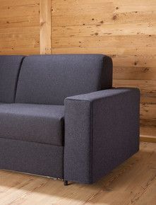 Ein graues Sofa mit breiten Armlehnen steht auf einem Holzboden vor einer Holzwand.