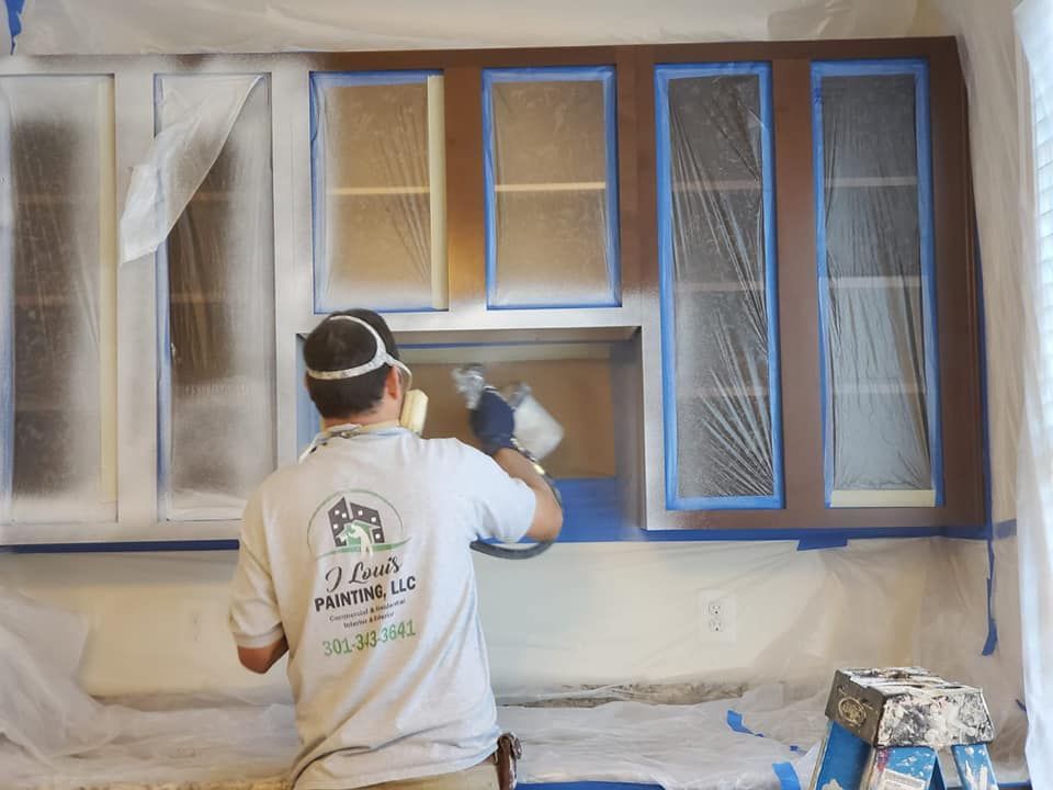 A worker spray-paints kitchen cabinets covered in plastic film and blue painter's tape.