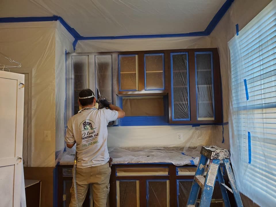 A person in a gray shirt spray-paints brown kitchen cabinets white in a room protected by plastic sheeting and blue tape.