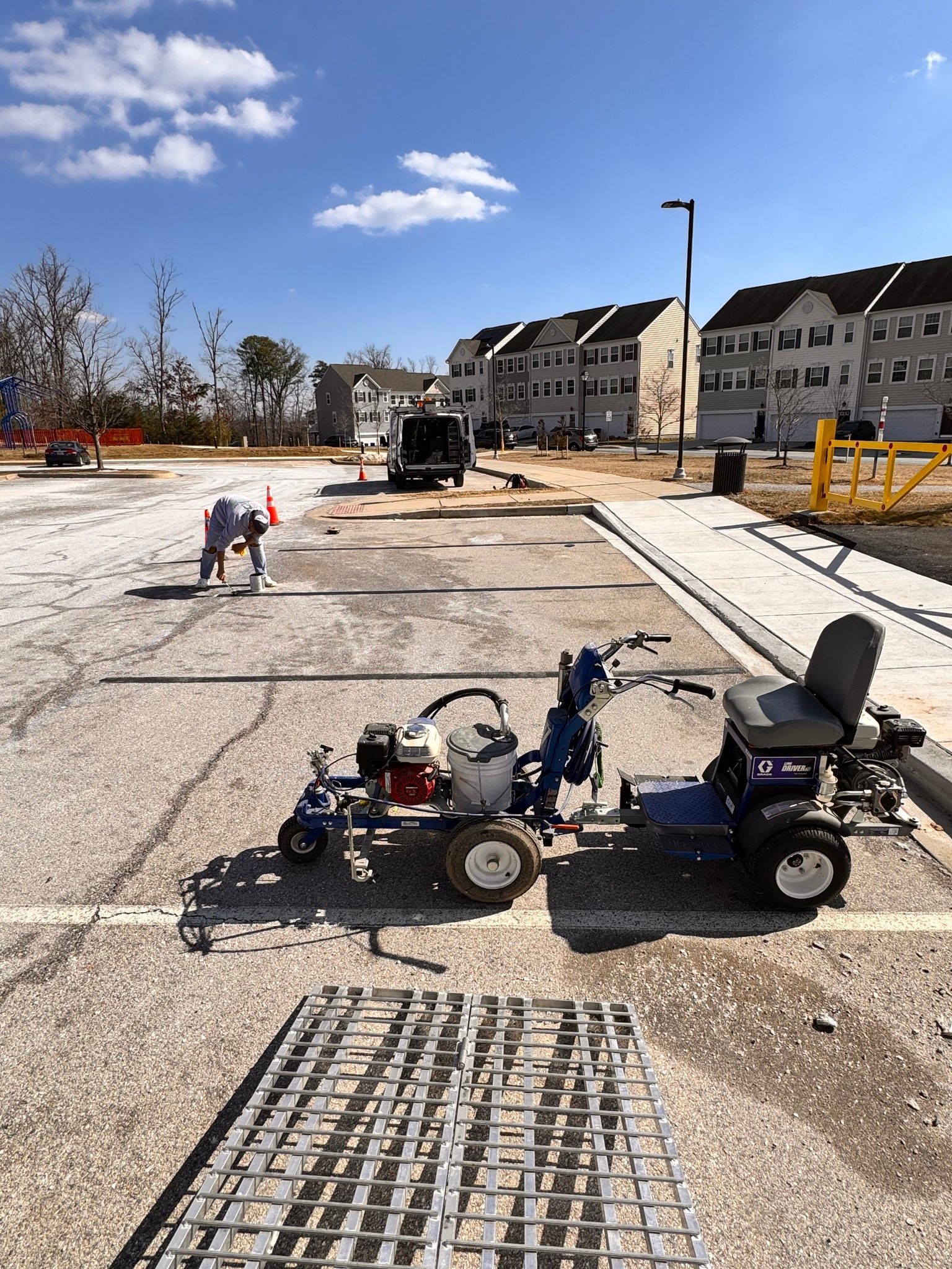 A worker uses a striping machine to paint parking lines on a gravel lot near residential buildings on a sunny day.