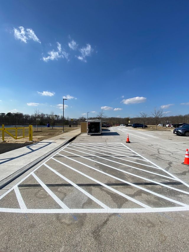A paved parking lot featuring a designated white-striped no-parking zone, with a trailer and cars visible in the distance.