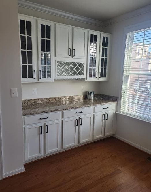 White kitchen cabinetry with glass doors, a wine rack, and granite countertops situated next to a window with blinds.