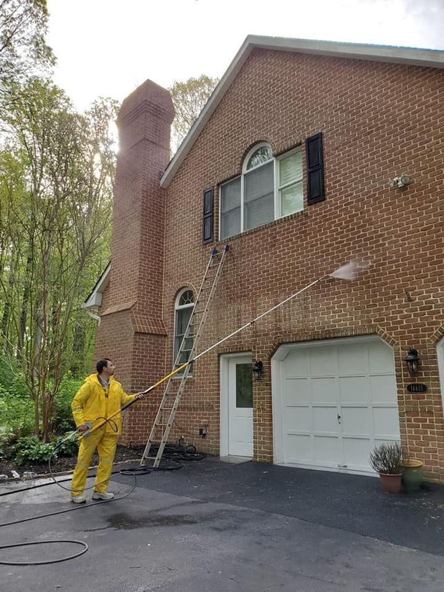 A person in a yellow rain suit uses a long pole to pressure wash the second-story exterior of a brick house.