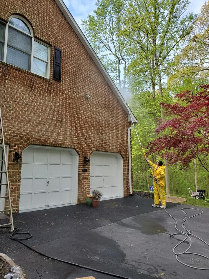 A person in a yellow rain suit pressure washes the exterior of a two-story brick house near a driveway and trees.