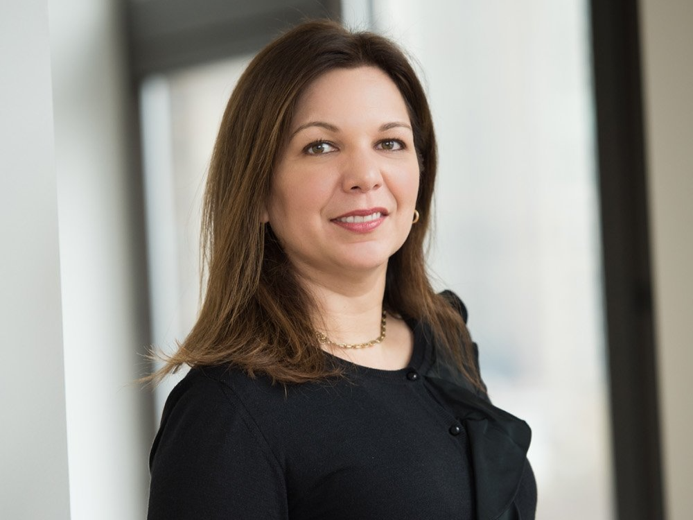 A woman in a black dress is standing in front of a window and smiling.