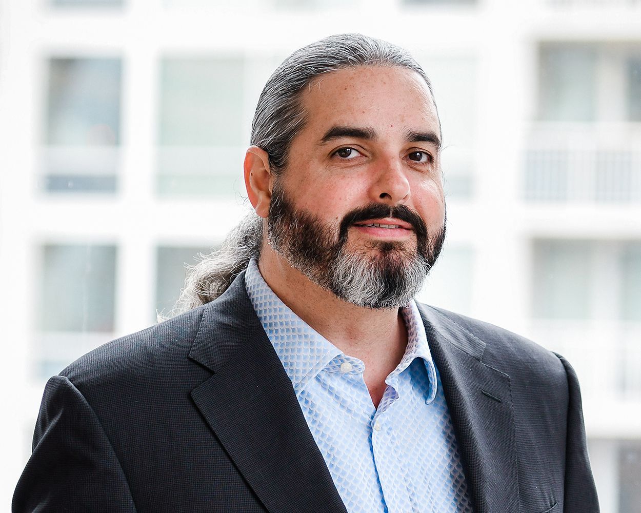 A man with a beard and long hair is wearing a suit and a blue shirt.