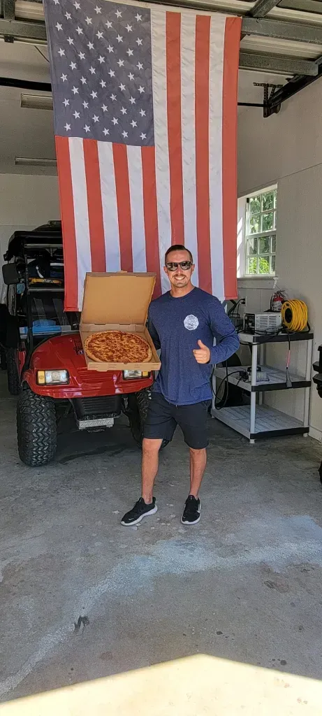 A man is holding a pizza in front of an american flag in a garage.
