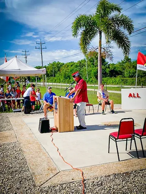 A man stands at a podium in front of a crowd with a sign that says all