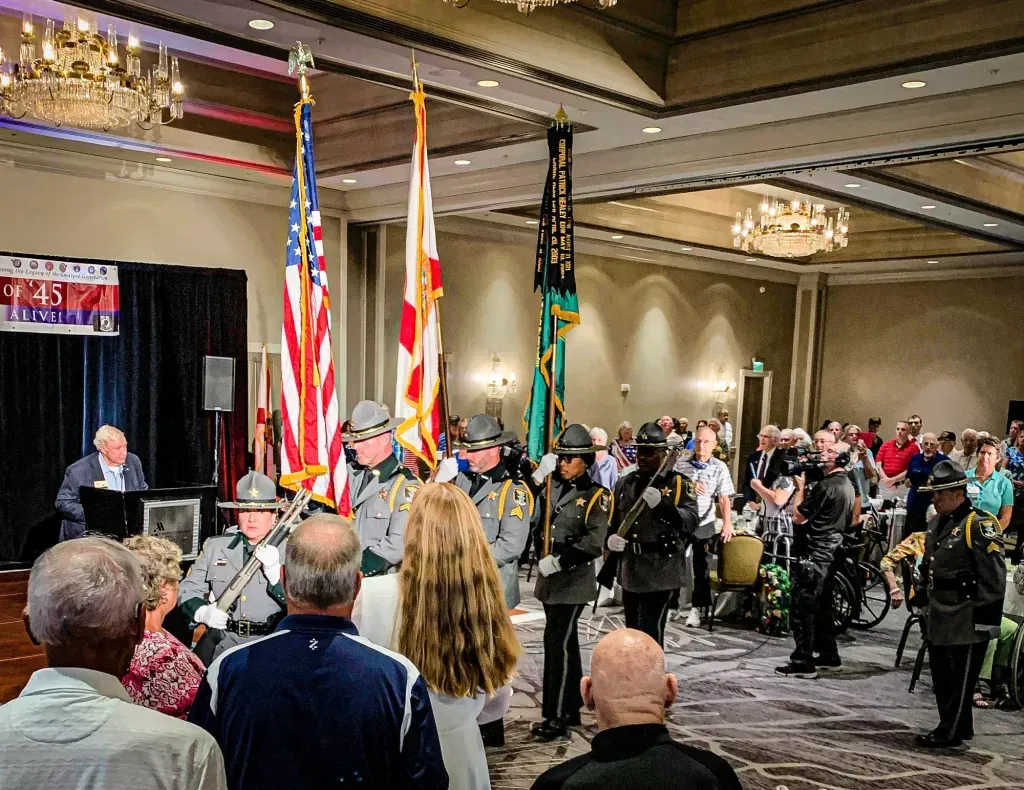 A group of people are standing in a large room watching a military parade.