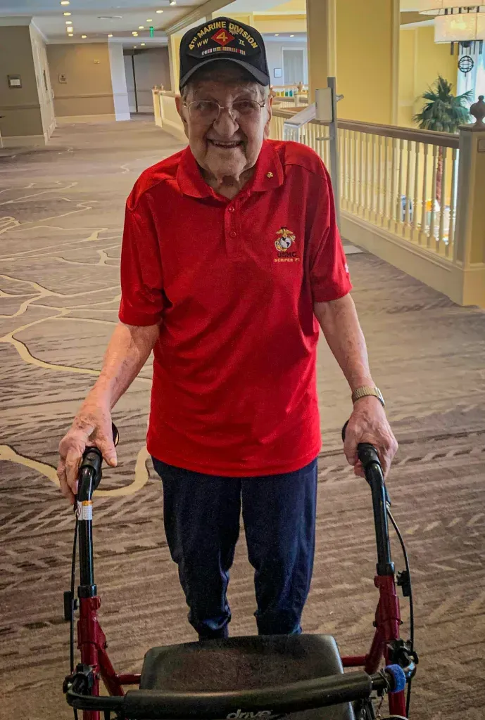 A man in a red shirt is walking with a walker in a hallway.