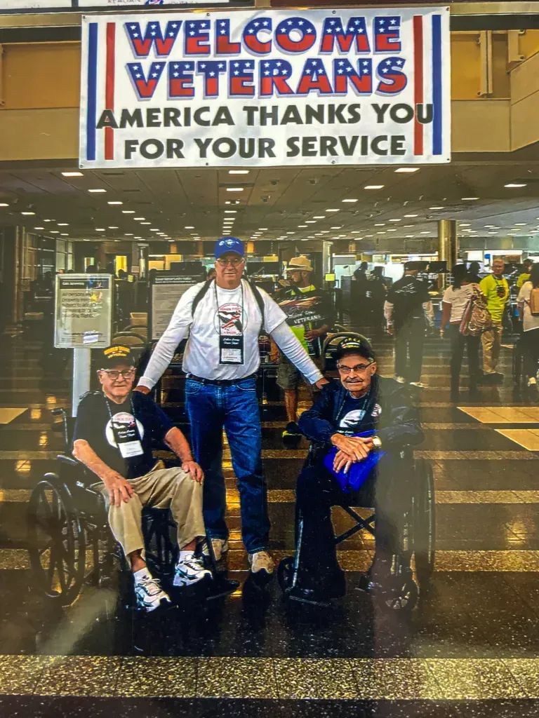 Three men are posing for a picture in front of a sign that says welcome veterans america thanks you for your service