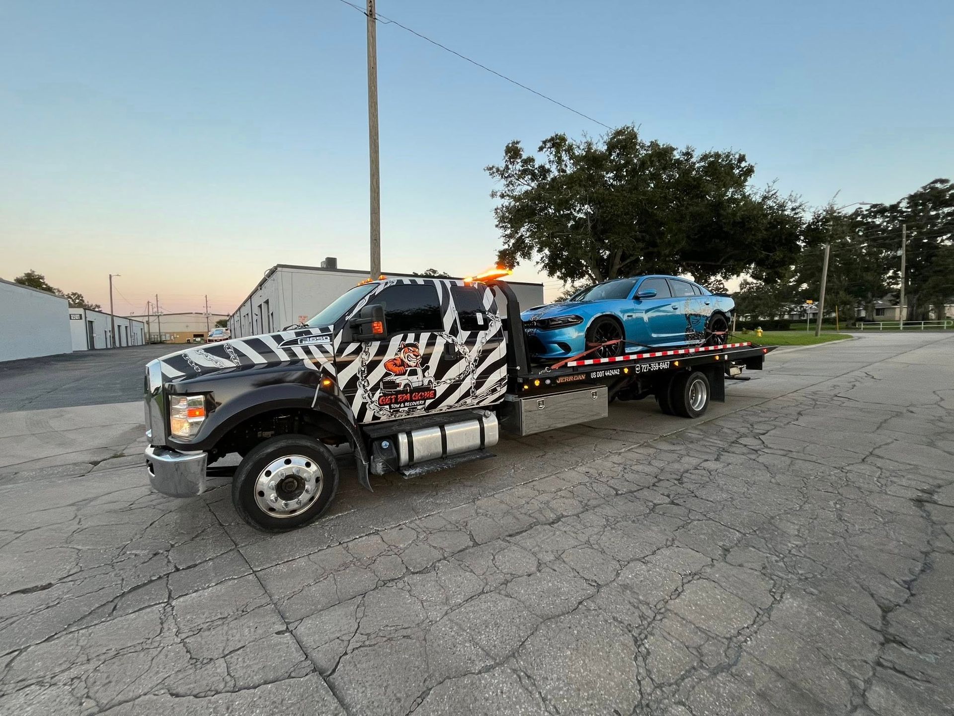 Tow truck with zebra stripes carrying a blue sports car on a cracked asphalt surface.