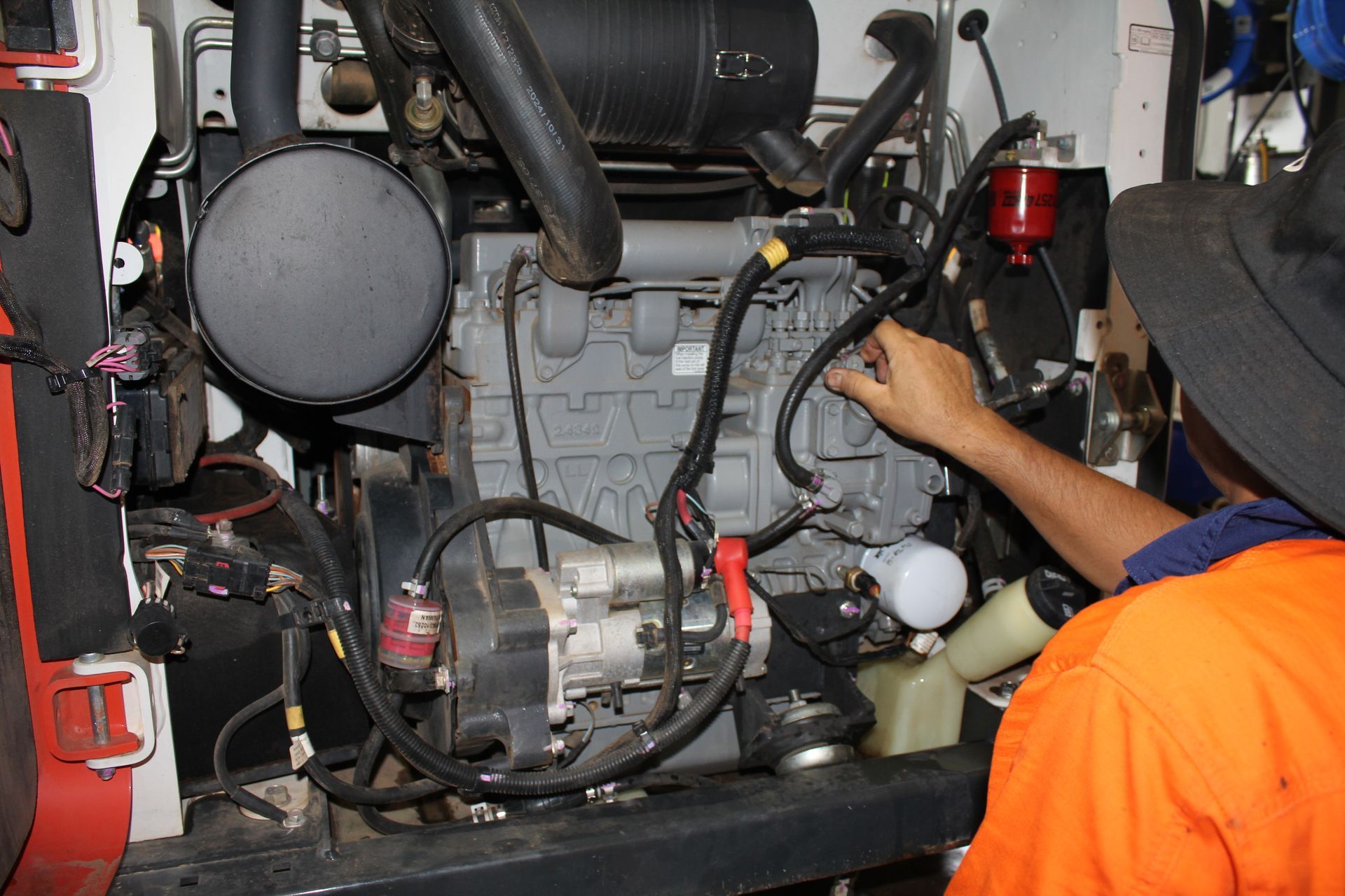 Mechanic working on an engine, reaching to a wire. Engine is gray, man wears orange shirt and hat — Forecast Machinery in Yarrawonga, NT